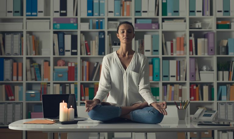 Relaxed woman practicing meditation at home at night, she is sitting in the lotus position on the office desk
